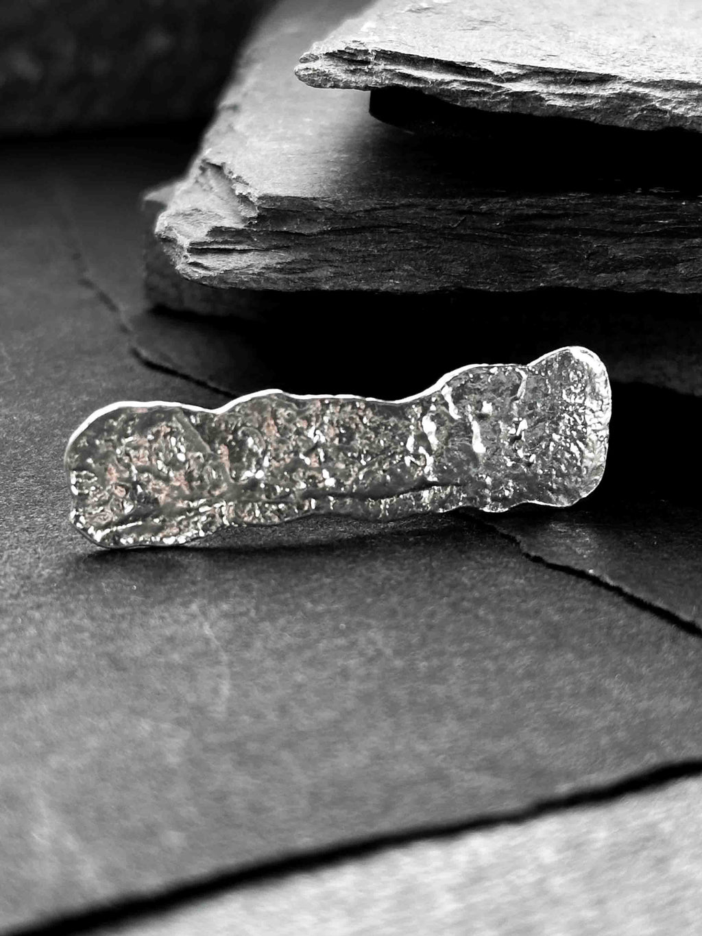 A hand wearing a large, rectangular textured silver ring with a ripped, organic edge and rippling molten texture.