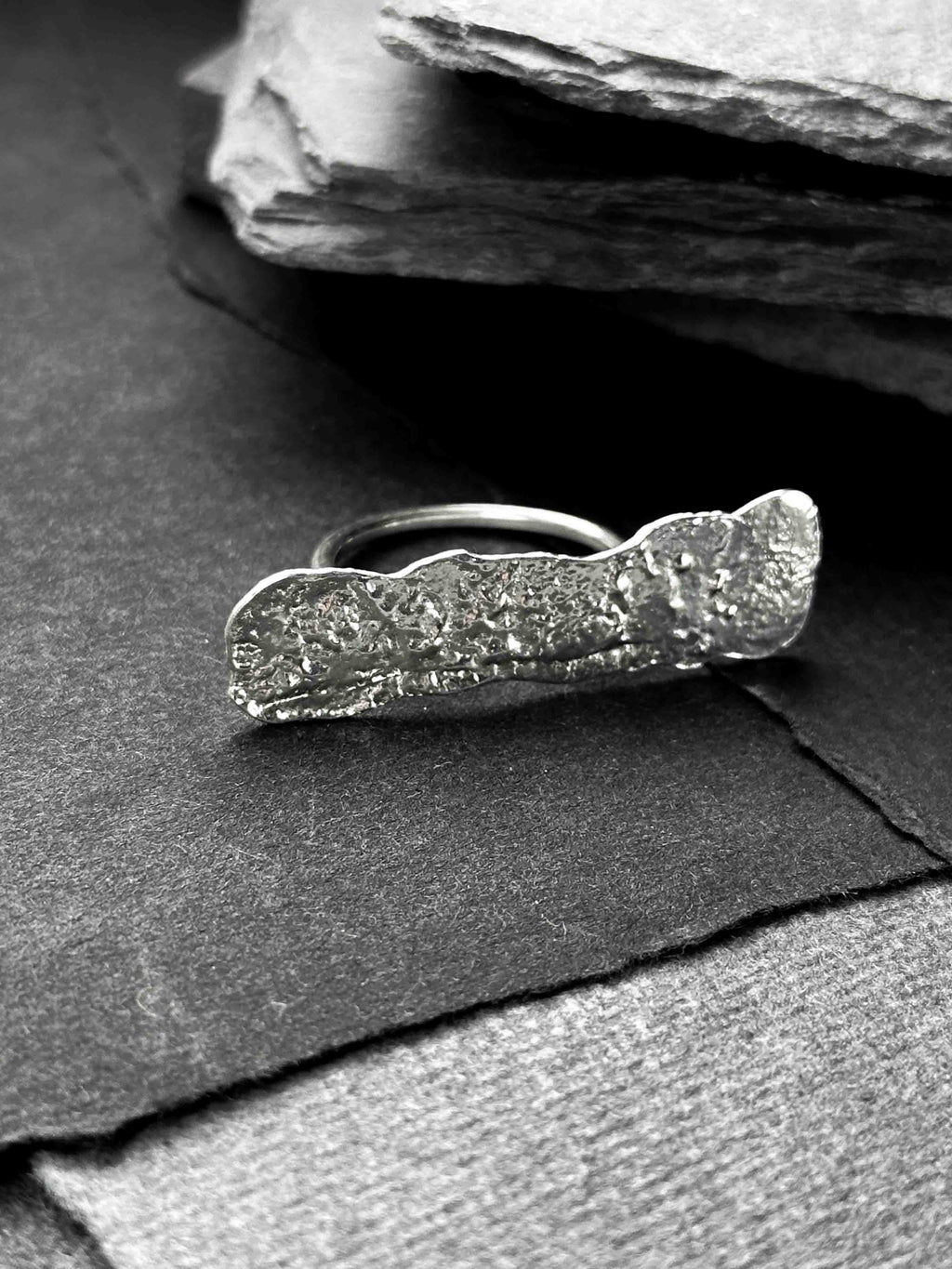 A large, rectangular textured silver ring with a ripped, organic edge and rippling molten texture.