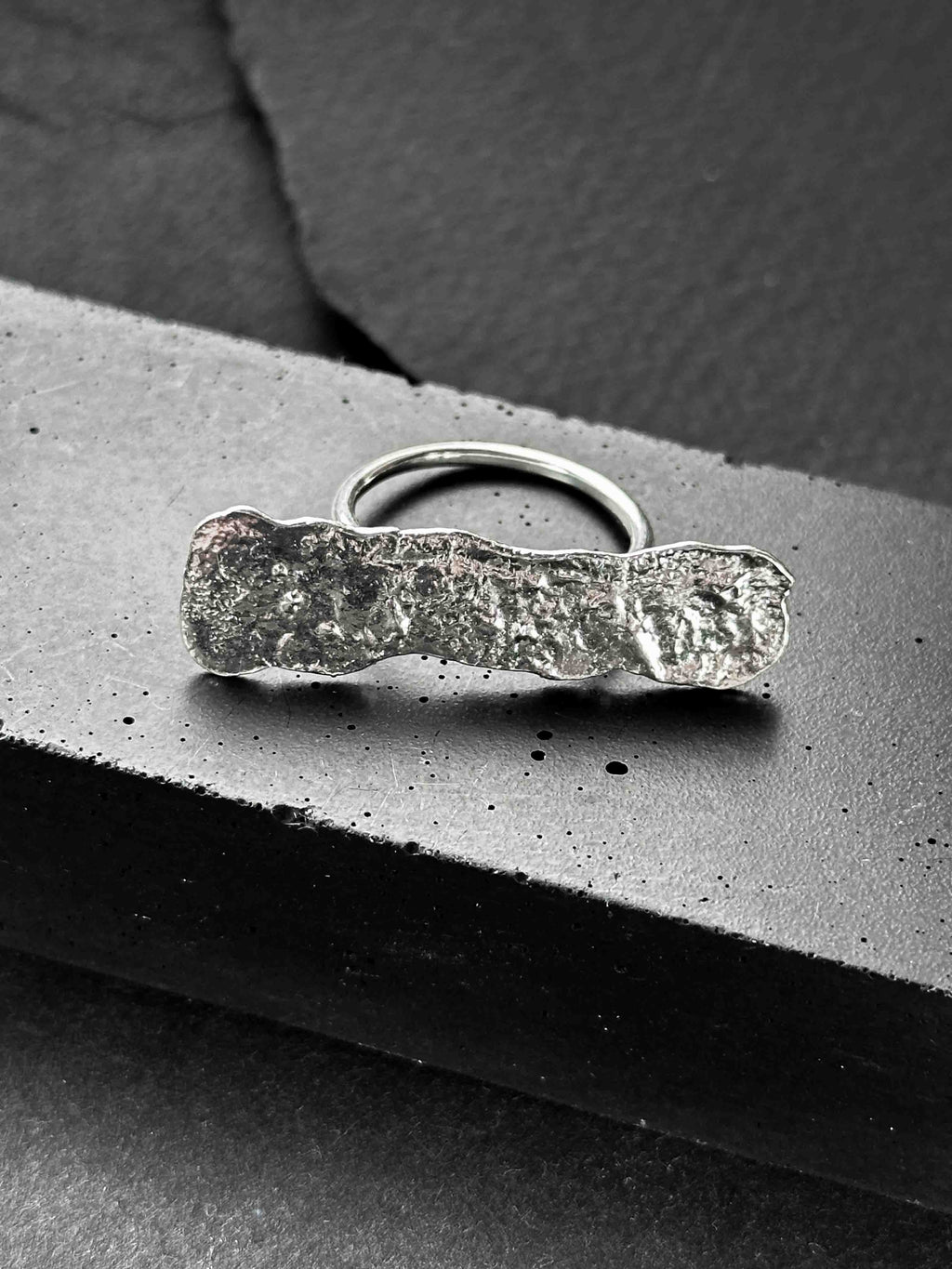 A large, rectangular textured silver ring with a ripped, organic edge and rippling molten texture.